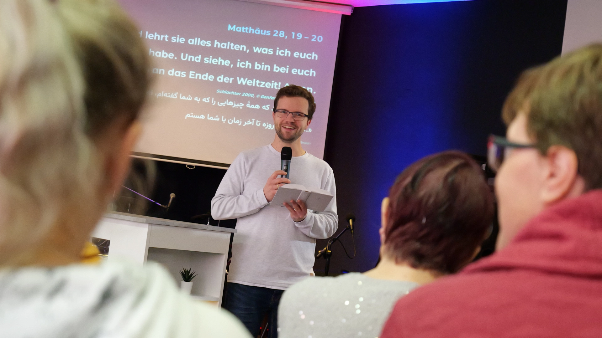 Benjamin Weis, Pastor der Elim Eichsfeld Gemeinde, hält ein Mikrofon und eine Bibel in der Hand und schaut zur Gemeinde lächelnd